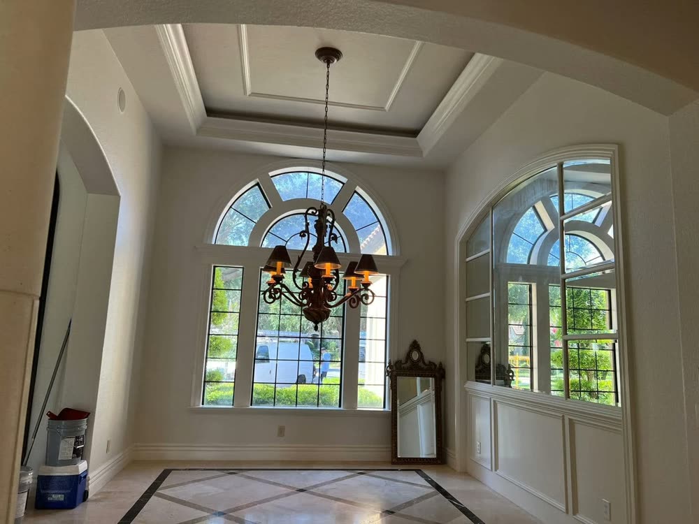 A room with a stepped tray ceiling, tall arched windows and a chandelier hanging on a long chain.