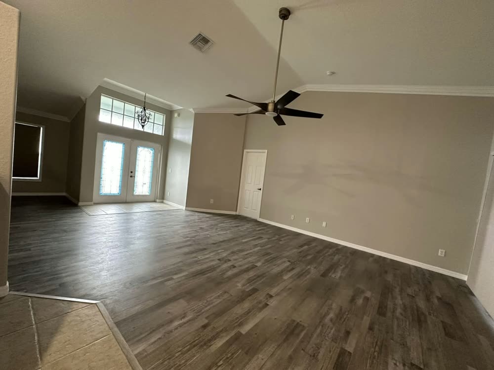 An empty living room with a vaulted ceiling, a ceiling fan and a glazed front door at the far end.
