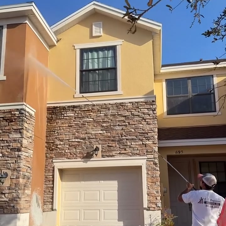 An SB Pro worker in a company shirt washing a two-storey townhome with a
                    long-reach wand, the spray arcing onto the pale upper wall above the
                    stonework at the base.